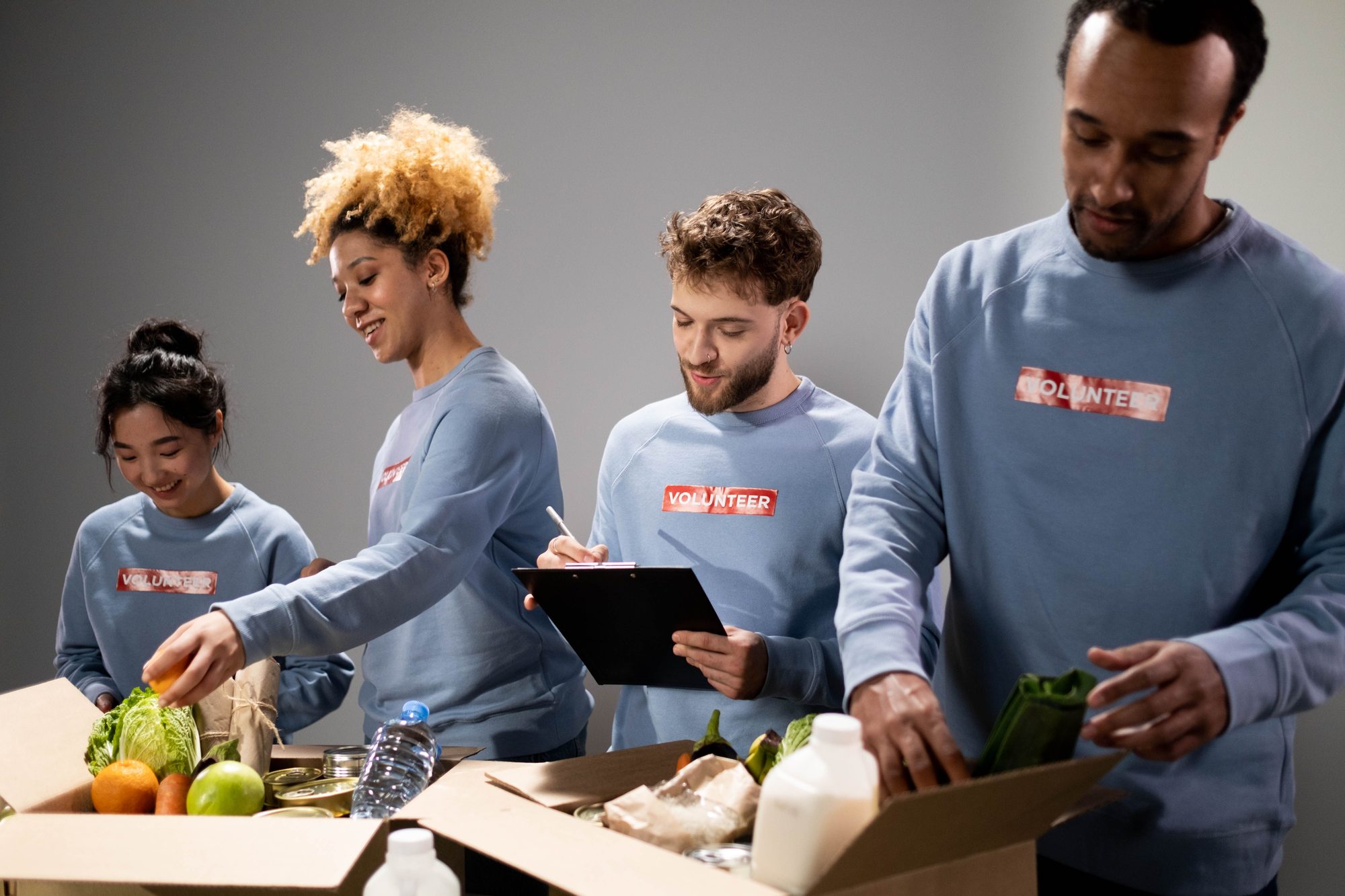 Four volunteers in matching sweatshirts sort fresh produce, canned goods, and bottled water into a food-donation box
