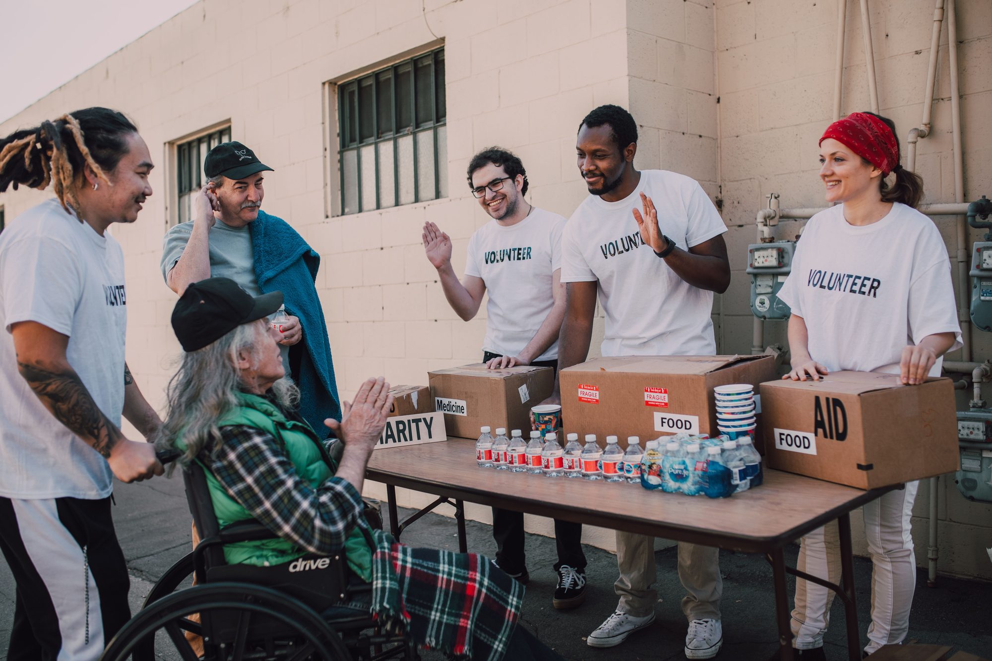 Volunteers in matching t-shirts warmly greet community members, including a man in a wheelchair, at an outdoor table stocked with food, water, and medical-aid boxes