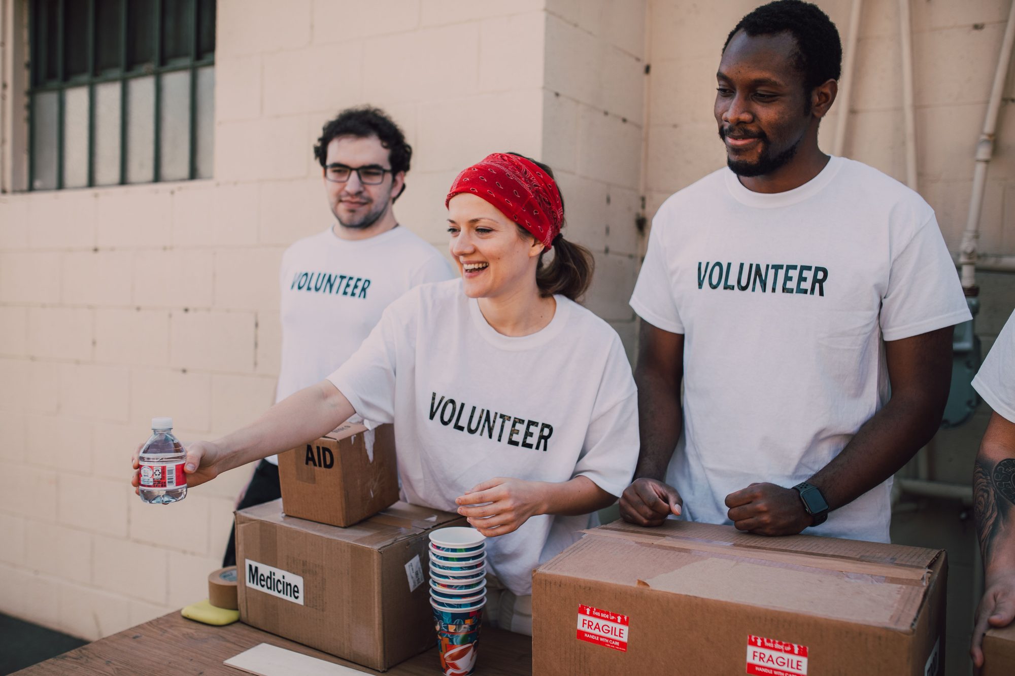 Three volunteers stand together at an outdoor aid station stocked with water bottles and supply boxes labeled Medicine and Aid